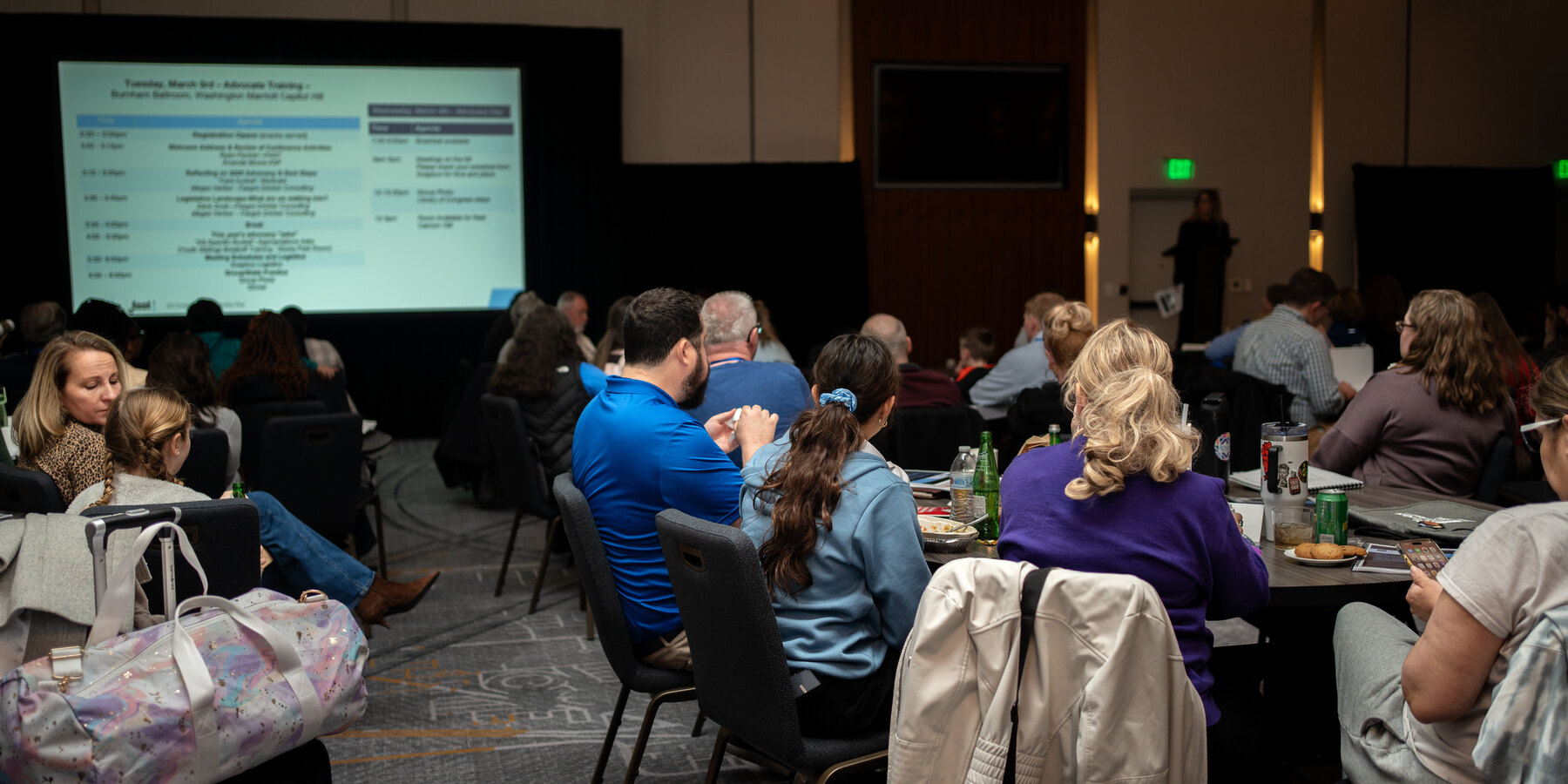 Attendees seated at tables reviewing the meeting agenda on screen